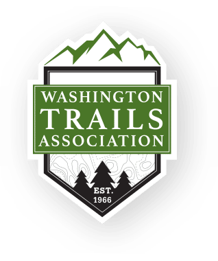 A white shield outlined in black, the top of the shield has an illustration of mountains and the bottom has pine trees. Washington Trails Association is written in a green bar across the center